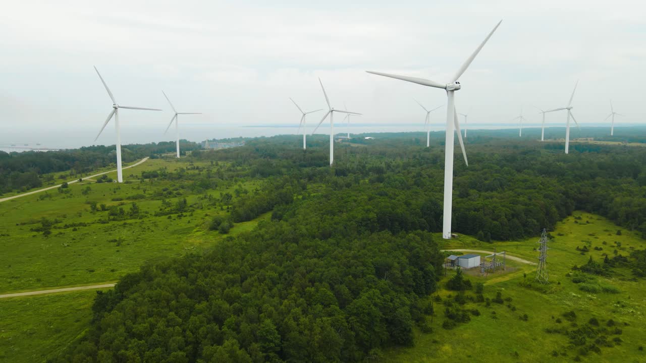 Aerial orbiting view of wind turbines in motion around greenery wooded area, electric windmills propeller rotating at the Paldiski peninsula wind farm. Clouded foggy sea coastline in the background