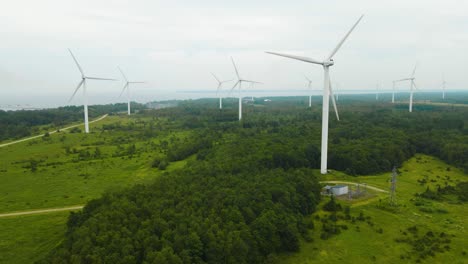 aerial orbiting view of wind turbines in motion around greenery wooded area, electric windmills propeller rotating at the paldiski peninsula wind farm. clouded foggy sea coastline in the background