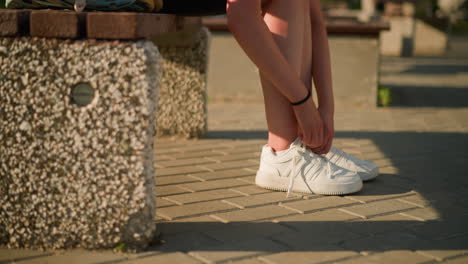 lower view of individual loosening sneaker shoelace while seated on bench, wearing white sneakers and black bangle on right hand, sunlight casts shadow on ground