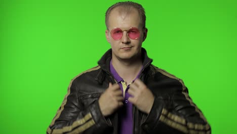 hombre rocker fresco en chaqueta de vaqueros marrón se pone gafas de sol rosas, mira a la cámara y aplaude sus manos