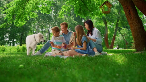 Happy-family-feed-white-dog-on-weekend-picnic.-People-rest-with-pet-in-park.