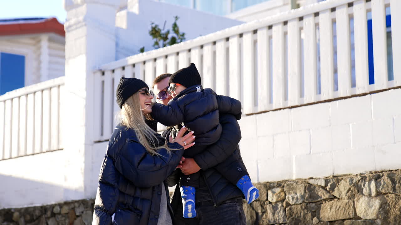 Family wearing black jackets and sunglasses in winter outdoors. Dad holds a baby and mom plays with her son.