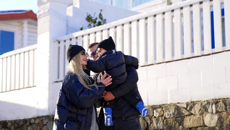 family wearing black jackets and sunglasses in winter outdoors. dad holds a baby and mom plays with her son.