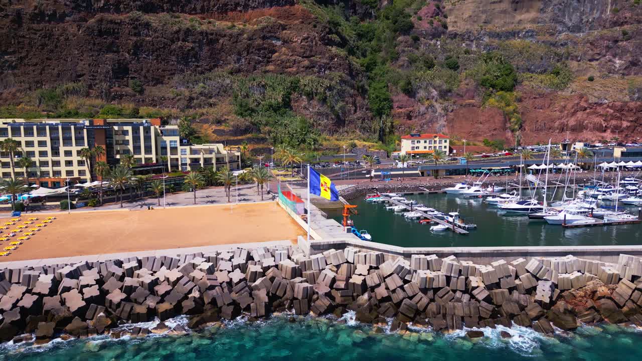 Aerial footage of the beach, marina and seawall in Calheta, Madeira, with flags, boats and coastal scenery. Ideal for travel, tourism, waterfront resorts and European destination themes