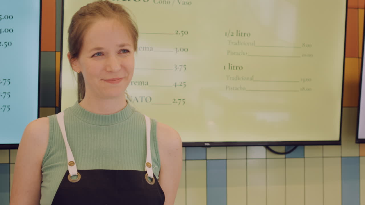 Medium shot of a young, light-eyed female ice cream shop employee handing out a sample of ice cream with a friendly and cordial expression.