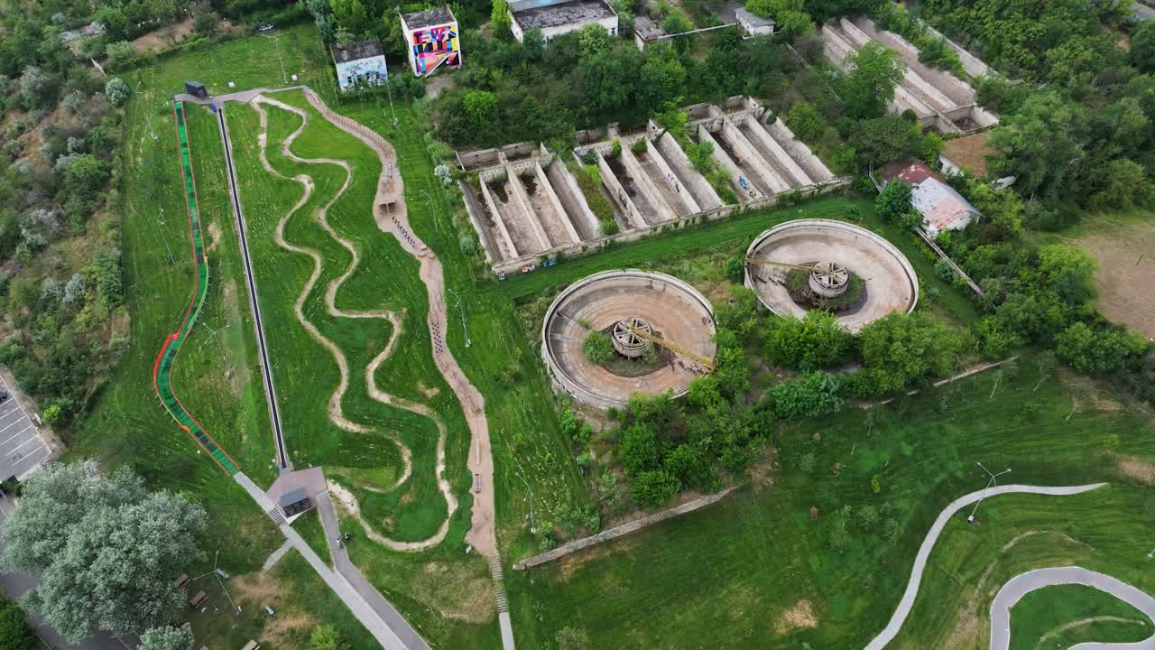 Abandoned wastewater treatment facility surrounded by dense overgrown vegetation. The decaying infrastructure includes empty sedimentation basins, circular clarifiers, aerial wide view