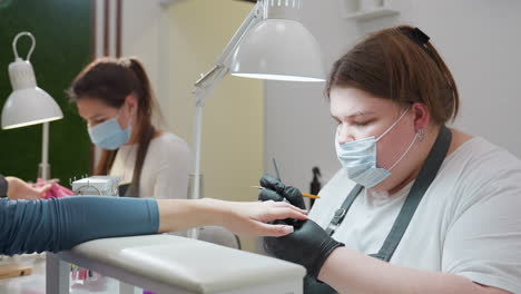 two nail technicians wearing face masks focus intently on client nails under bright lamps one files nails in background while other polishes with precision in foreground, capturing professionalism