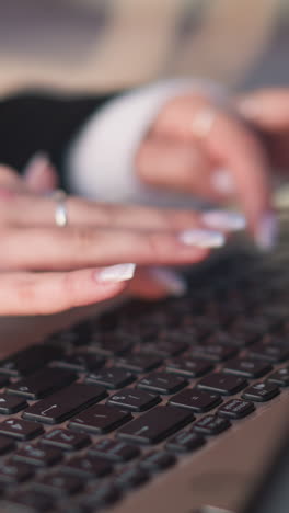 close-up of hands with polished nails and delicate rings typing on laptop keyboard outdoors, placed on wooden table with blurred background