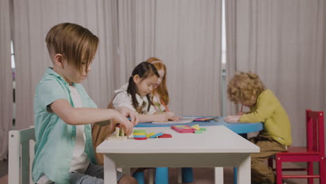 Niño-Pequeño-En-Una-Escuela-Montessori-Jugando-Con-Formas-Apiladas-Mientras-Sus-Compañeros-De-Clase-Dibujan