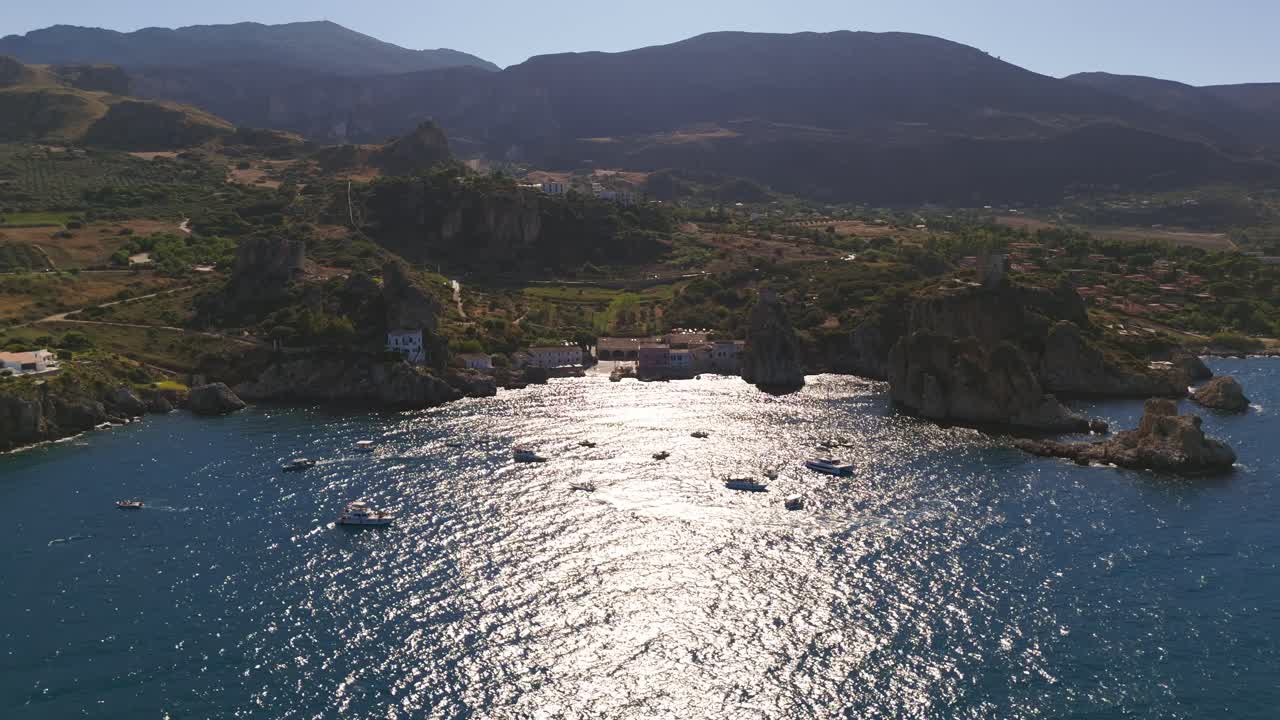 Aerial drone shot pulling back over the turquoise waters of Tonnara di Scopello, Sicily, Italy, gradually revealing numerous boats floating in the calm sea during a bright and warm Italian summer day