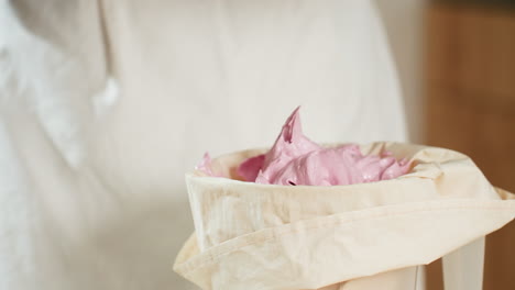 close-up of fluffy pink dough in piping bag as baker scrapes and prepares it for use in cake decoration or pastry filling, with soft and smooth texture visible in cozy kitchen environment