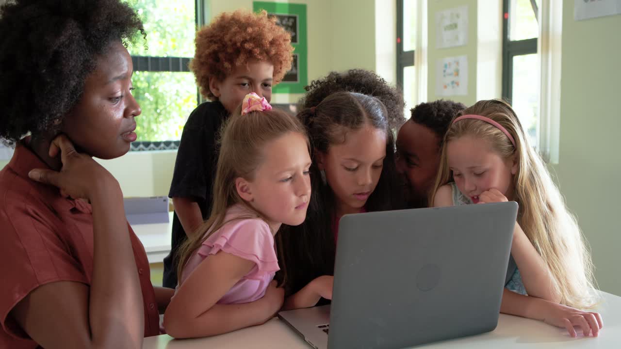African American teacher pointing guiding kids leaning and seeing laptop in classroom on white desk