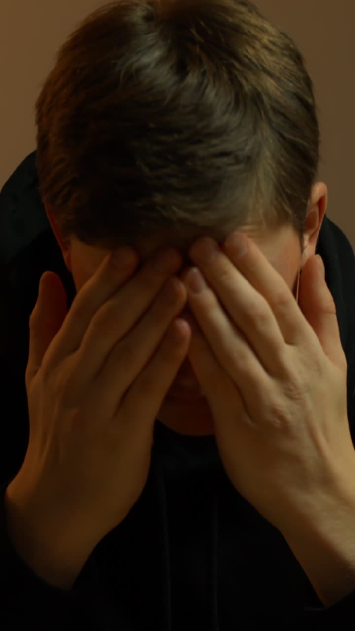Close-up de um jovem cobrindo o rosto, mostrando estresse e frustração em um ambiente interno sob luz quente: um momento emocional forte para temas de saúde mental ou burnout
