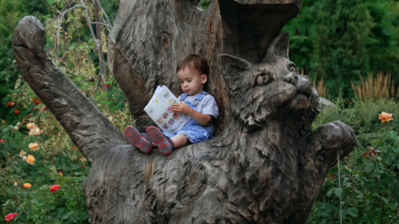 Small Caucasian kid sits on a big wooden cat. Lovely child looking through a baby magazine.