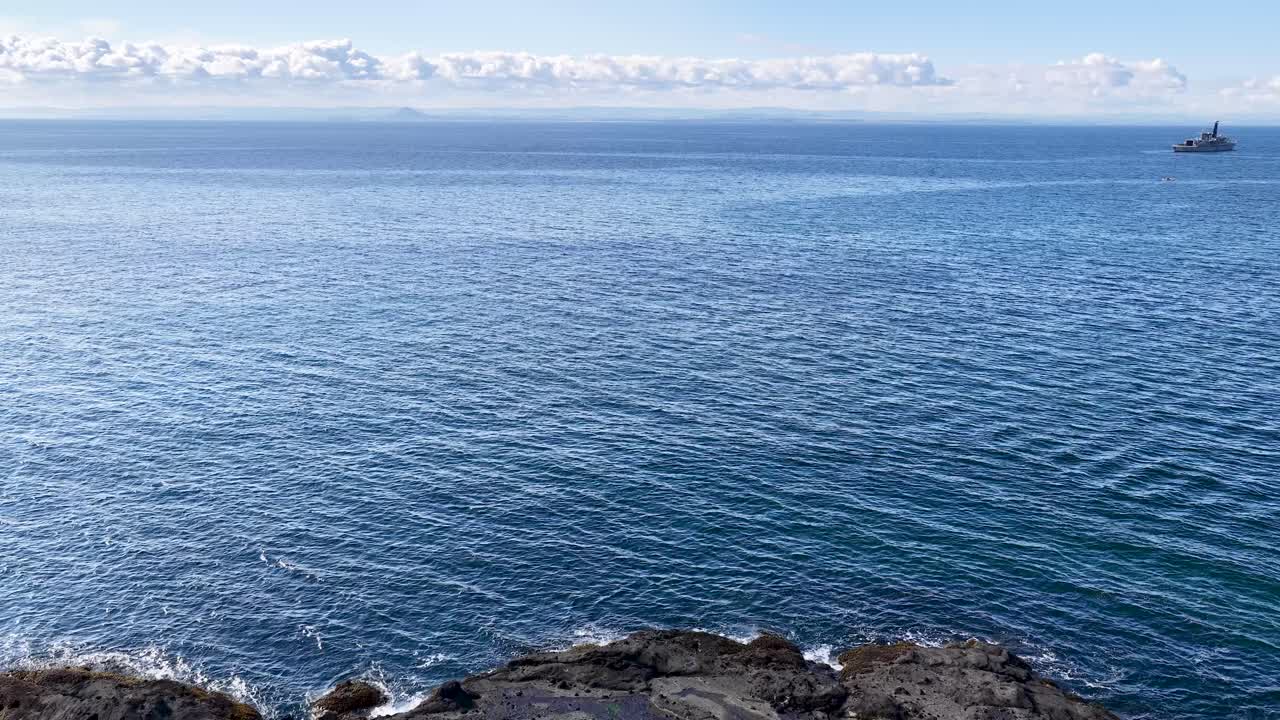 Daytime camera pan reveals rocky shoreline, historic building, flagpole, and tranquil ocean horizon