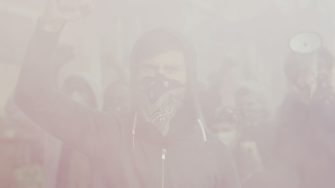 vista de cerca de un joven caucásico con capucha y bufanda en la cara protestando y gritando alrededor de humo y gas en la calle con otras personas