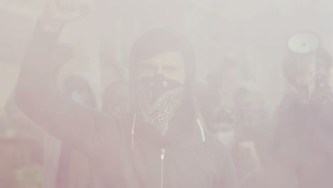 close-up view of caucasian young man in hood and scarf on his on face protesting and yelling around smoke and gas in the street with other people