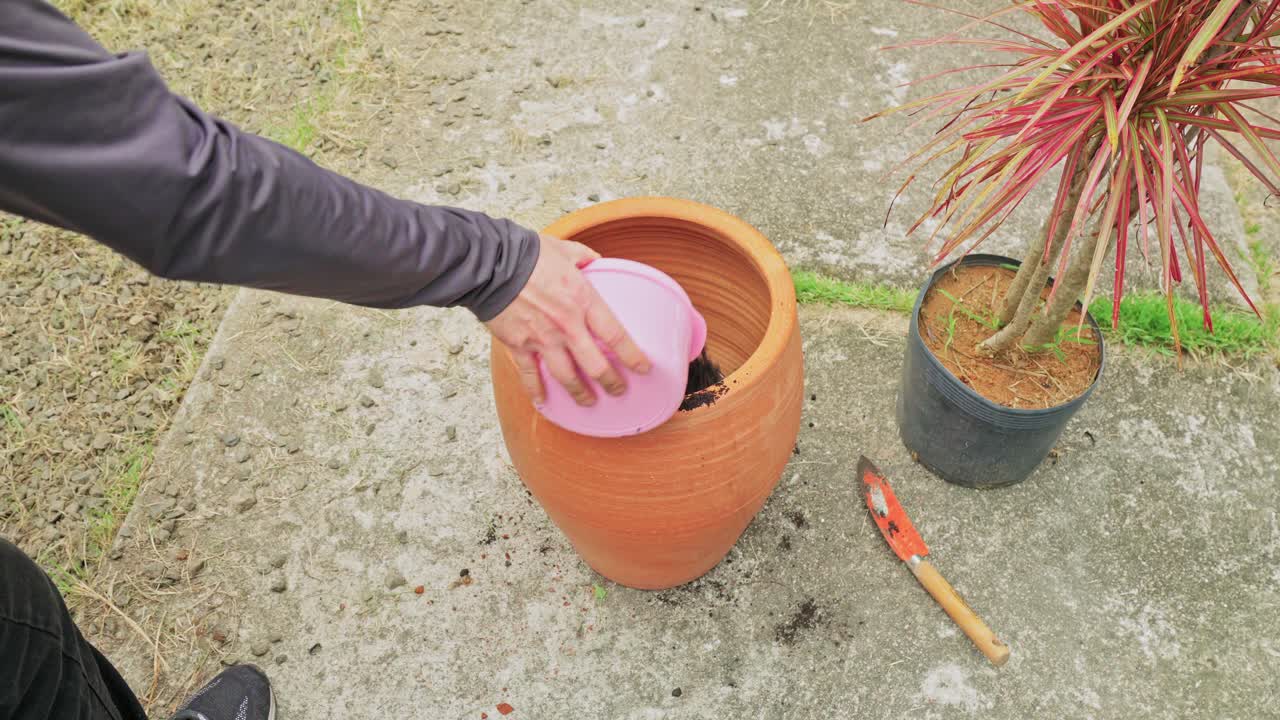 High angle shot of a person using a small trowel to add potting soil around a newly placed plant inside a large round terracotta planter in a backyard