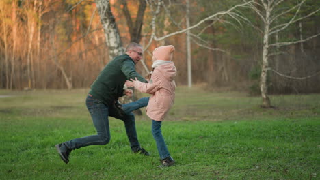 a man in a green jacket and a little girl in a pink jacket, both wearing blue jeans, are seen joyfully jumping and playing together outdoors on a lush green grassy field