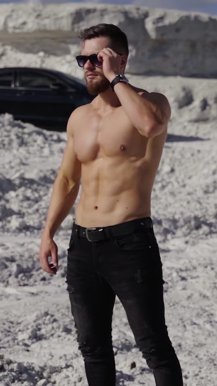 Athlete with muscular body in white mountains. Handsome sportsman without shirt in sunglasses stands on canyon on black car background. Vertical video