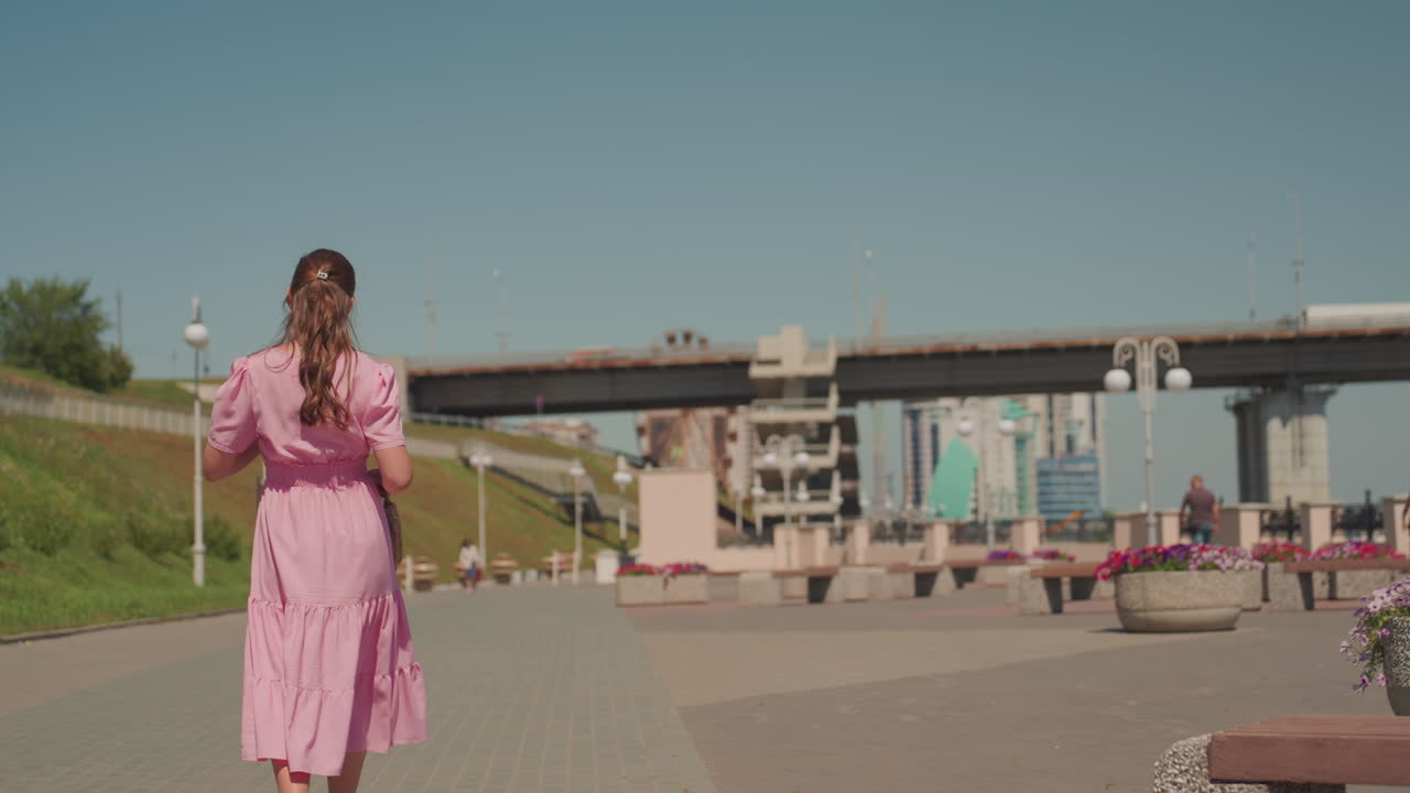 Donna caucasica in abito rosa che passeggia lungo una soleggiata passeggiata urbana vicino a un ponte e al lungomare, si ferma vicino a fioriere vivaci mentre controlla il telefono, atmosfera estiva rilassata con toni cinematografici pastello