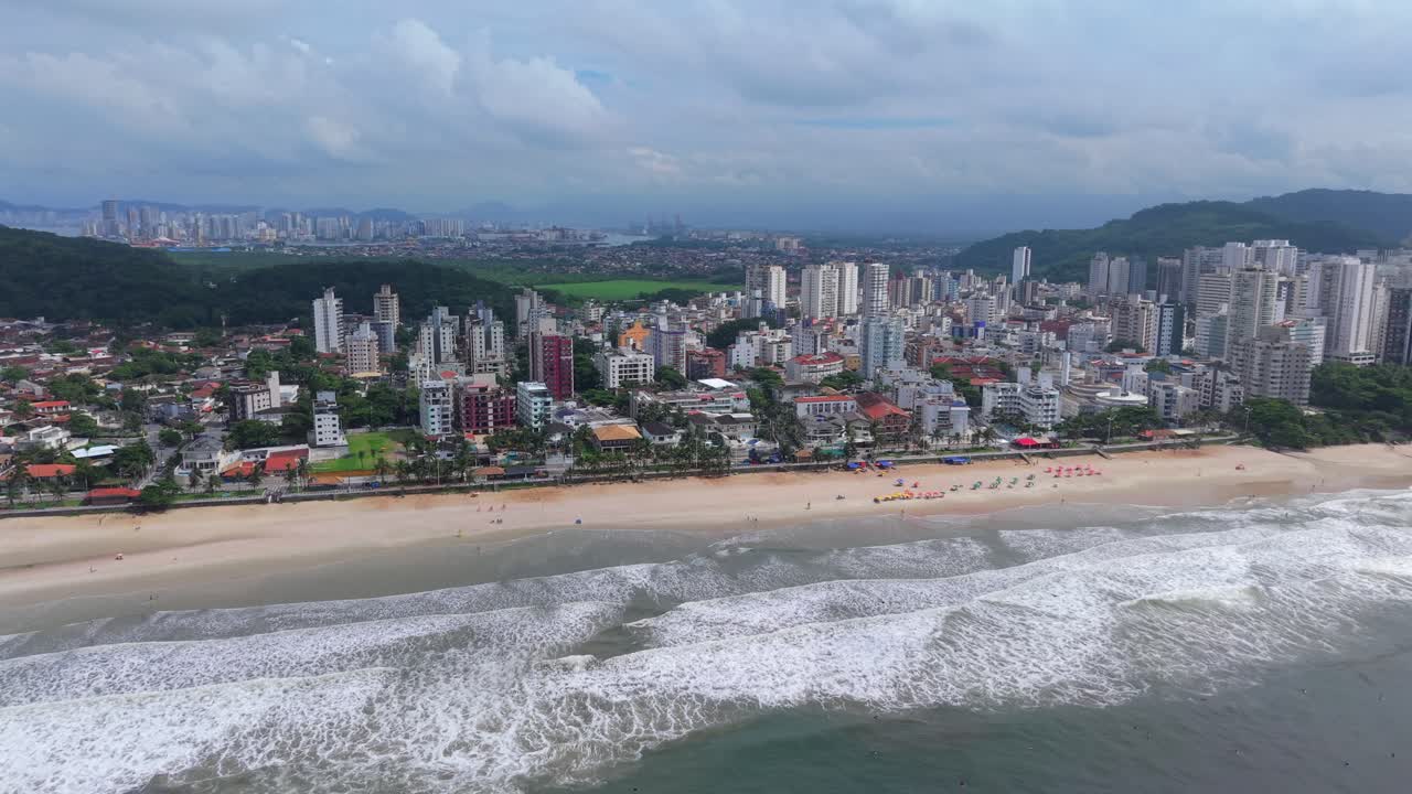 Praia do Tombo Guarujássa, São Paulossa, Brasiliassa, hiekkarantoineen, Atlantin valtameren aallokkoineen ja tiheine asuinalueen siluetteineen, jota tukevat Serra do Mar -vuoret, rannikkokaupunkimaisema, hidas drone-dolly-sisäänajo.