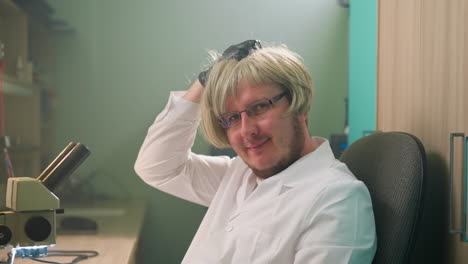 una vista en primer plano de un técnico con una bata de laboratorio blanca y una peluca rubia, rascándose juguetón la cabeza con una mano de guante negro mientras está sentado en un laboratorio junto a un microscopio