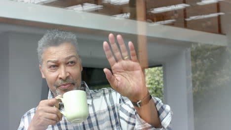 hombre bi-racial senior feliz saludando por la ventana y bebiendo café en casa, espacio de copia, cámara lenta