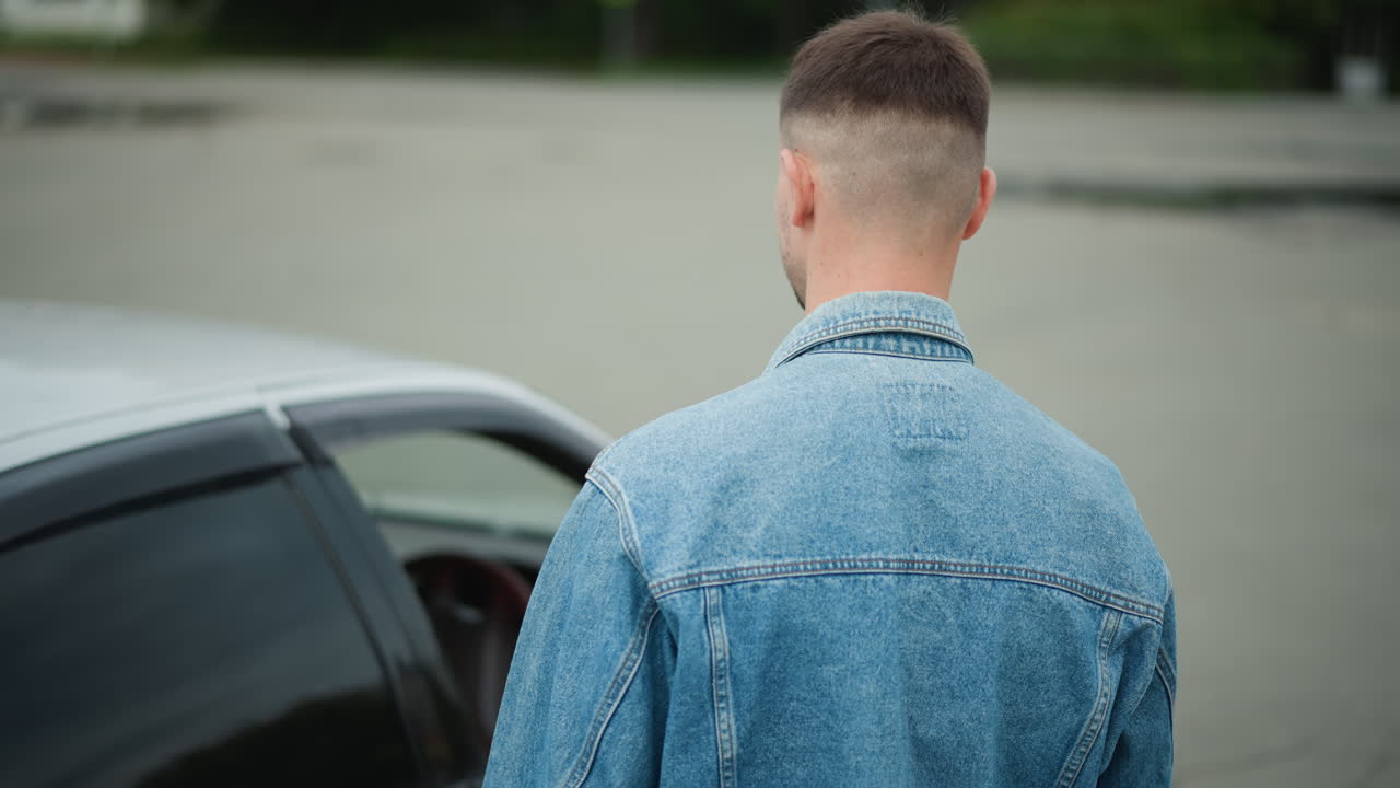 Motorista verifica o carro cautelosamente nas proximidades, indivíduo vestindo jeans observa e inspeciona o automóvel estacionado com cuidado, homem de jaqueta jeans examina cuidadosamente o carro estacionado em um cenário urbano deserto