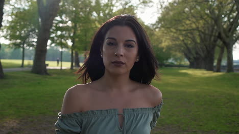 slow motion portrait of a gorgeous hispanic latino young woman looking at the camera and show emotions from serious to smiling and laughing with a beautiful british park in the background