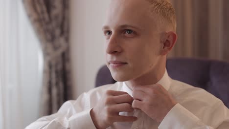 Stylish-man-in-jacket-fastens-buttons,-buttoning-white-shirt-preparing-to-go-out-near-window