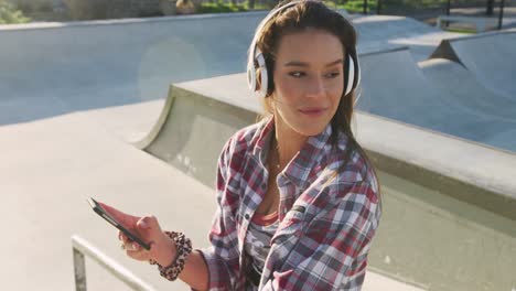 Smiling-caucasian-woman-wearing-headphones,-using-smartphone-and-waving-at-a-skatepark