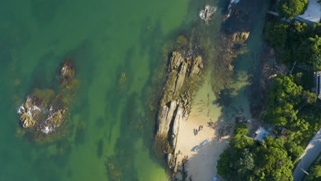 Aerial-top-view-panning-over-the-shore-of-a-brazilian-paradise-beach-with-white-sand,-emerald-clear-water,-boats-and-rocks