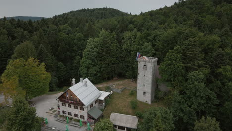 Drohnenschuss-Von-Oben-Auf-Einen-Gipfel-Des-Berges-Mirna-Gora,-Wo-Sich-Eine-Berghütte-Und-Ein-Turm-Mit-Einer-Slowenischen-Flagge-Befinden,-Die-Im-Wind-Flattert