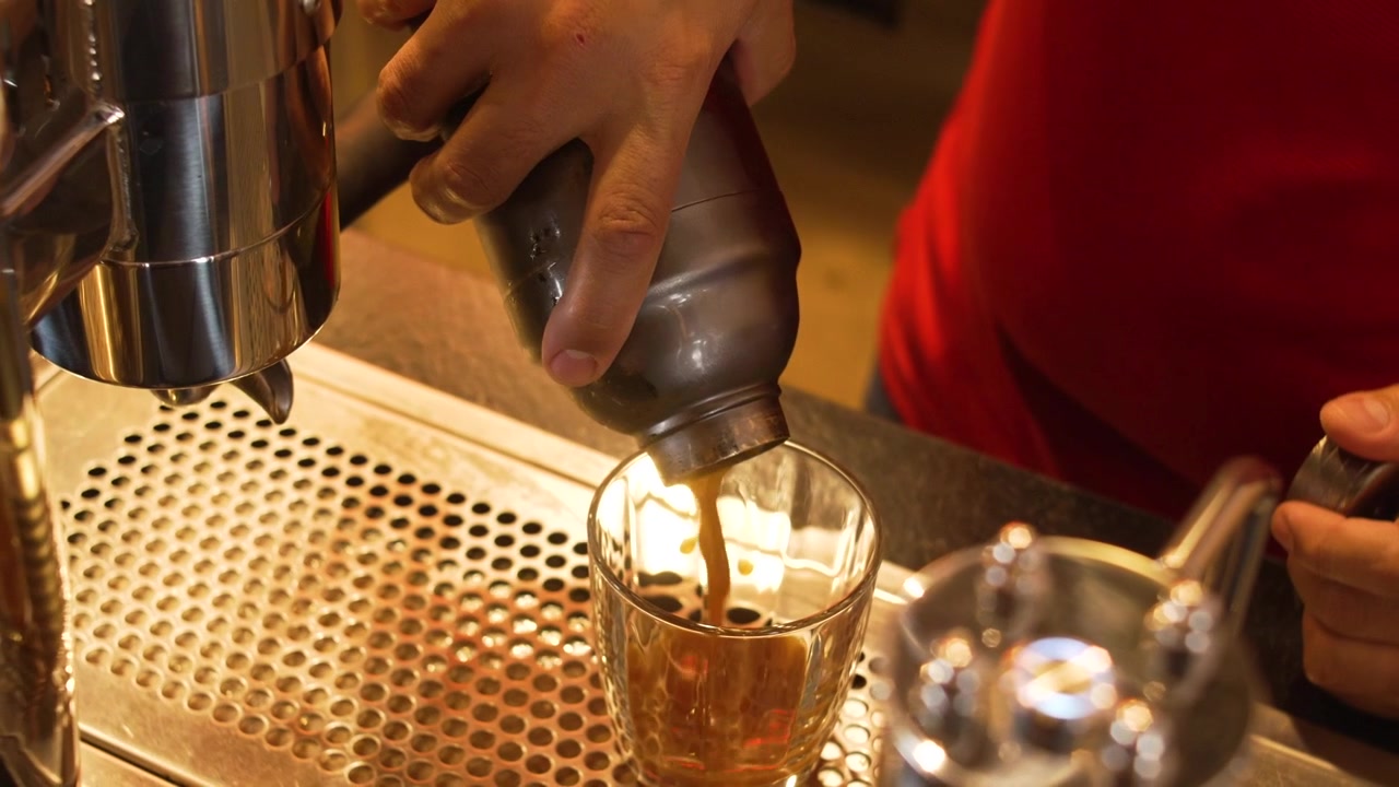 Nhân viên pha chế mặc đồng phục đỏ rót tách espresso tươi vào ly đá tại máy pha cà phê chuyên nghiệp, cảnh hành động tại quán cà phê đặc biệt