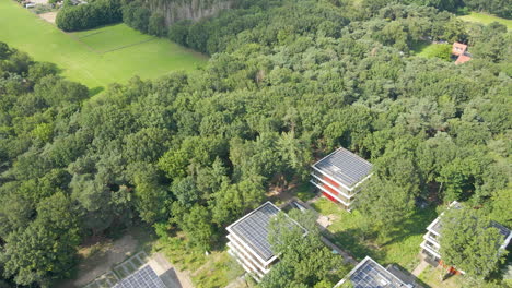 aerial of small office buildings with solar panels on rooftop in a green forest and a parking lot providing shadow to cars with solar panels