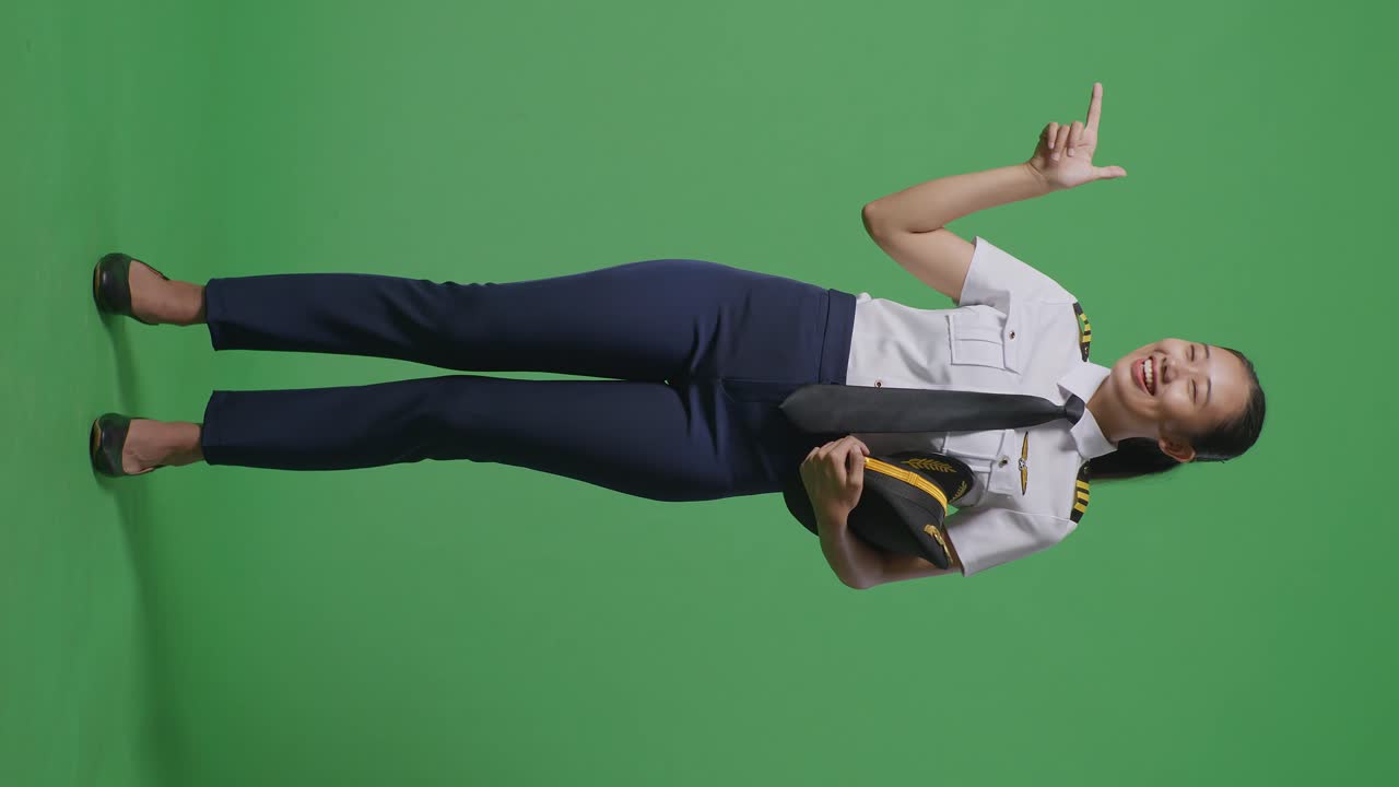 녹색 스크린 배경 스튜디오에 서 있는 동안 웃고 을 가리키는 아시아 여성 조종사의 몸이 가득 차 있습니다.
