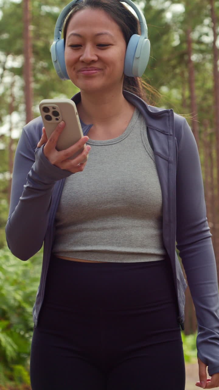 Video Verticale di Donna con Cuffie Wireless che si Allena all'Aperto, Ascoltando Musica o Podcast in Streaming dal Telefono Cellulare, Correndo Lungo un Sentiero nel Bosco e Indossando Abbigliamento Sportivo, Ripreso in Tempo Reale