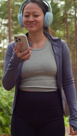 vertical video of woman wearing wireless headphones exercising doing work out outdoors streaming music or podcast from mobile phone running along track in forest wearing sports clothing shot in real time