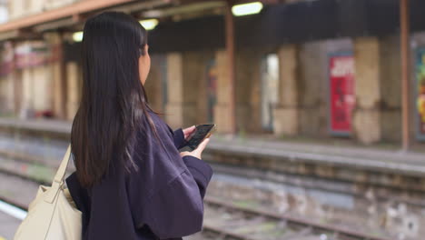 rear view of young woman waiting on railway station platform for train looking at mobile phone