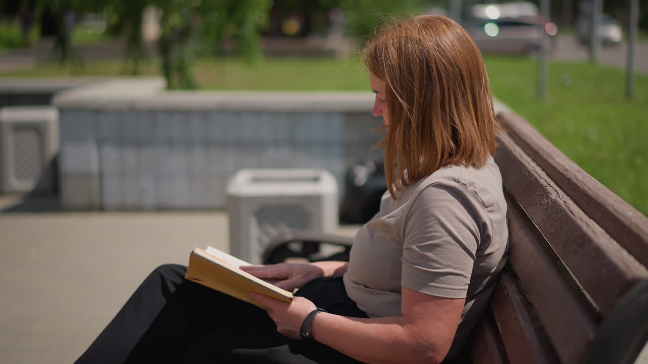 Donna seduta su una panchina di legno che legge un libro all'aperto in una giornata di sole, sfogliando le pagine con attenzione e un'espressione serena mentre delle auto sfocate passano sullo sfondo
