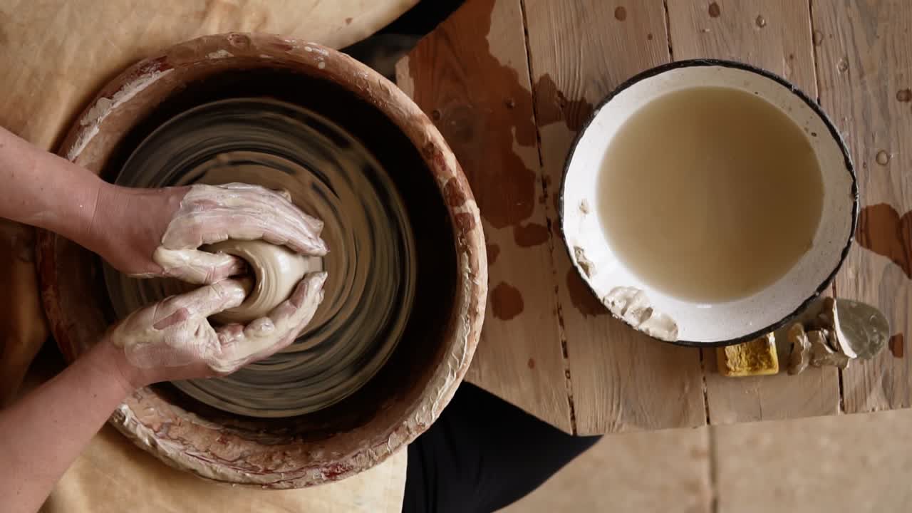 Close-up top view of potter's hands with working with wet clay on a pottery wheel making a clay product in a workshop. Unrecognizable female person formong vase, bowl with a water on a table