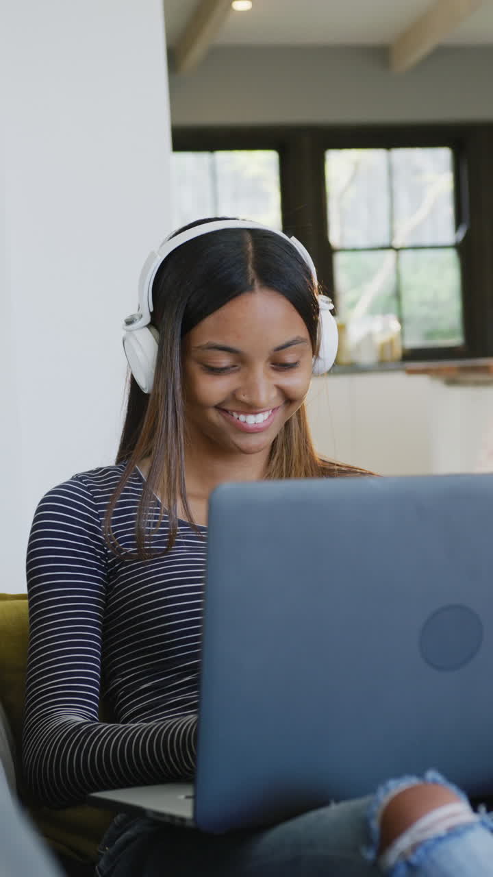 Vertical video of happy biracial teenager girl wearing headphones and using laptop, slow motion