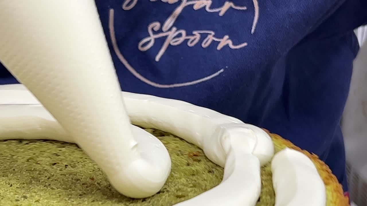 Primo piano estremo di un pasticcere professionista che fa girare lentamente il supporto girevole, spremendo la crema al burro di albumi montati dalla sac à poche per decorare il bordo della base di pan di Spagna
