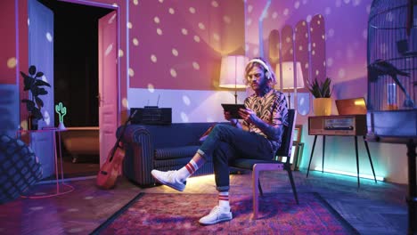 Young-stylish-caucasian-man-with-long-hair-in-headphones-and-glasses-sitting-on-a-chair-and-using-a-tablet-in-a-retro-party-at-home