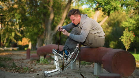 young man with light brown hair seated on reddish-brown log in park, wearing light gray sweatshirt and khaki pants, focused on fastening spring stilts with concentrated or mildly frustrated expression