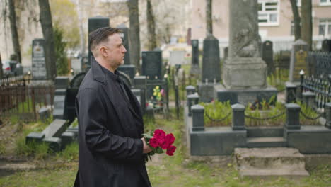 side view of man in black raincoat and suit holding roses while walking in a graveyard 1