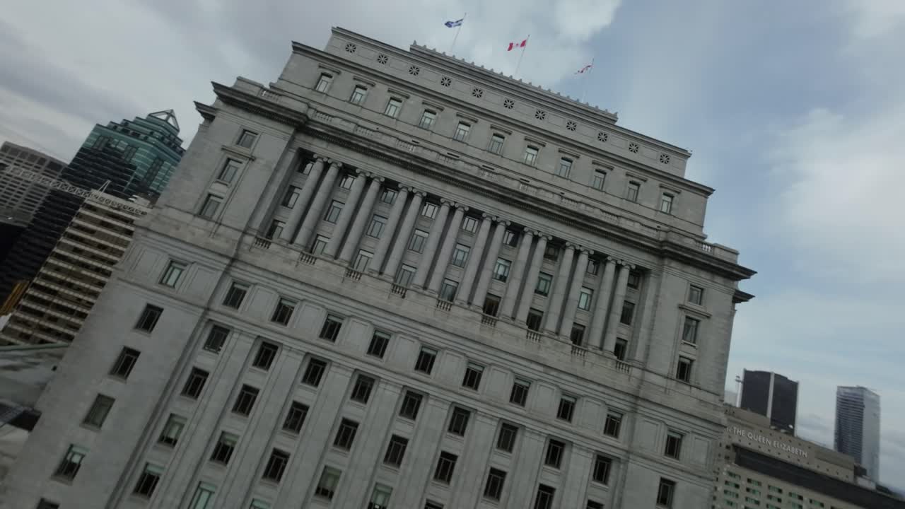 FPV drone pushing towards iconic Sun Life Building's top, highlighting its grandeur and architectural and flying flags on top in downtown Montreal's Square Dorchester.
