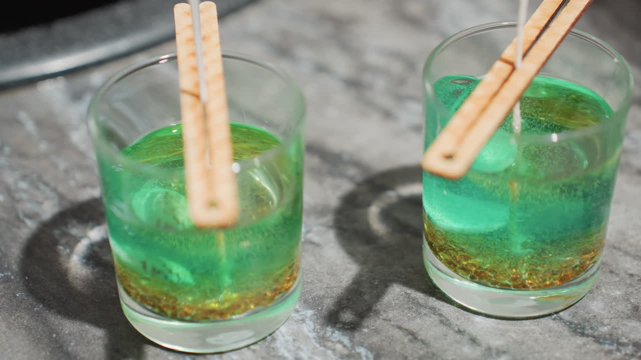 Three glass cups filled with layered green and brown melted wax mixture, each with a centered wick held upright by wooden clips placed across cup rims, set on smooth gray kitchen counter