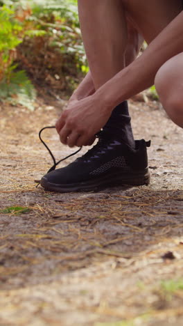 Vídeo-Vertical-De-Un-Hombre-Atándose-Los-Cordones-De-Una-Zapatilla-Deportiva-Antes-De-Hacer-Ejercicio-Corriendo-Por-Una-Pista-A-Través-De-Un-Bosque,-Filmado-En-Tiempo-Real-1